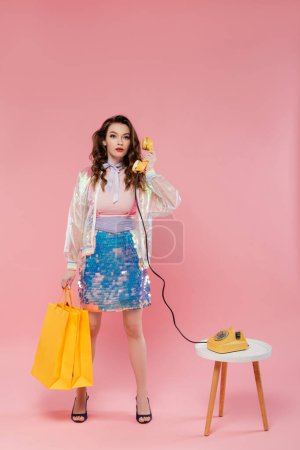 schöne junge Frau mit Einkaufstaschen, am Retro-Telefon sprechend, wie eine Puppe auf rosa Hintergrund stehend, Konzeptfotografie, Puppenpose, Modell im Rock mit Pailletten und transparenter Jacke 