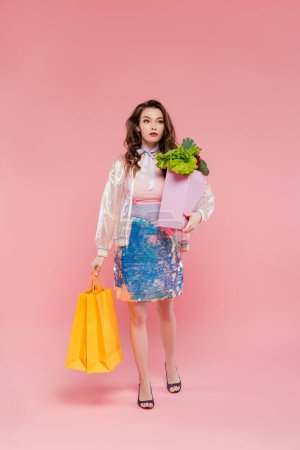 hermosa joven que lleva bolsas de compras y bolsa de comestibles, de pie sobre fondo rosa, fotografía conceptual, deberes en el hogar, alimentos y verduras, concepto de ama de casa 