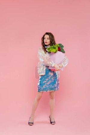 Photo for Housewife concept, attractive young woman carrying grocery bag with vegetables, model with wavy hair standing on pink background, conceptual photography, home duties, stylish wife - Royalty Free Image