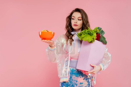concept de femme au foyer, attrayant jeune femme portant sac d'épicerie avec légumes et bol avec flocons de maïs, modèle avec cheveux ondulés sur fond rose, photographie conceptuelle, tâches à la maison, petit déjeuner 