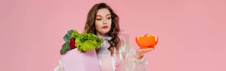 housewife concept, attractive young woman carrying grocery bag with vegetables and bowl with corn flakes, model with wavy hair on pink background, conceptual photography, home duties, banner