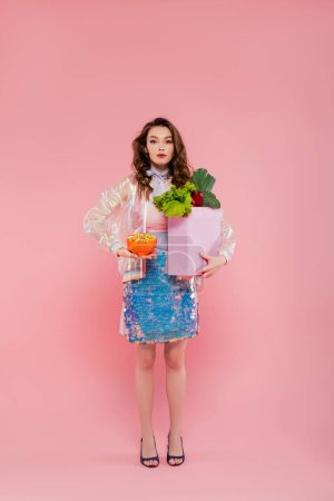 Hausfrauenkonzept, puppenhaft, attraktive junge Frau mit Gemüsetasche, Modell mit welligem Haar auf rosa Hintergrund, konzeptionelle Fotografie, häusliche Pflichten, stilvolle Ehefrau 
