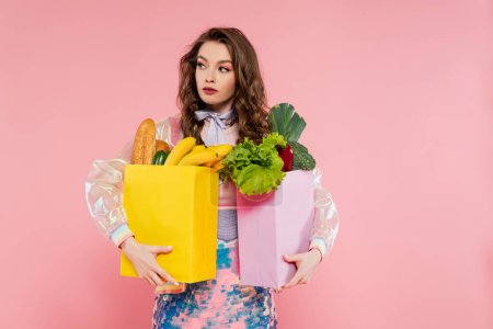 Hausfrauenkonzept, attraktive junge Frau, die Einkaufstüten mit Gemüse und Bananen trägt, Model mit welligem Haar auf rosa Hintergrund, konzeptionelle Fotografie, häusliche Pflichten, stilvolle Ehefrau 