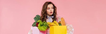 Hausfrauenkonzept, attraktive junge Frau, die Einkaufstüten mit Gemüse und Bananen trägt, Model mit welligem Haar auf rosa Hintergrund, konzeptionelle Fotografie, häusliche Pflichten, stilvolle Ehefrau, Banner 