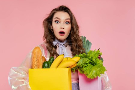 Hausfrauenkonzept, schockierte junge Frau, die Einkaufstüten mit Gemüse und Bananen trägt, Model mit welligem Haar auf rosa Hintergrund, konzeptionelle Fotografie, häusliche Pflichten, stilvolle Ehefrau 