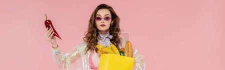 housewife concept, attractive young woman in sunglasses carrying paper bag with groceries and holding red pepper, posing like a doll on pink background, conceptual photography, banner 