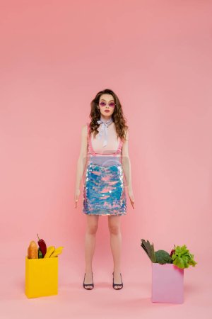 Hausfrauenkonzept, attraktive junge Frau mit Sonnenbrille, die wie eine Puppe neben Papiertüten mit Lebensmitteln posiert, brünettes Model mit welligem Haar auf rosa Hintergrund, konzeptionelle Fotografie, häusliche Pflichten
