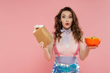 Hausfrauenkonzept, schockierte junge Frau posiert wie eine Puppe, hält Essen zum Mitnehmen und eine Schüssel mit Cornflakes, rosa Hintergrund, konzeptionelle Fotografie, häusliche Pflichten, emotionale 