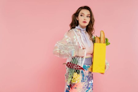 housewife concept, beautiful young woman holding reusable mesh bag with groceries, stylish wife doing daily house duties, standing on pink background, looking away, role play 