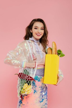 concept de femme au foyer, heureuse jeune femme tenant sac en filet réutilisable avec épicerie, femme élégante faisant des tâches quotidiennes de la maison, debout sur fond rose, regardant la caméra, jeu de rôle 