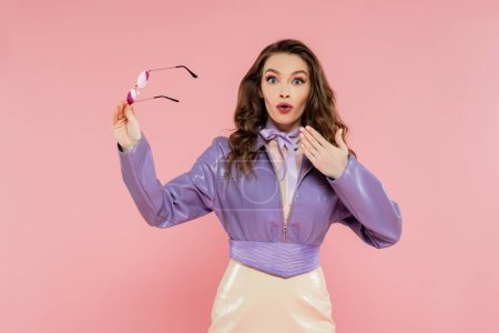 glamour, surprised young woman gesturing and looking at camera, holding sunglasses, fashionable outfit, model in purple jacket and skirt standing on pink background, studio shot, acting like a doll 