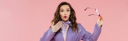 glamour, surprised young woman gesturing and looking at camera, holding sunglasses, trendy outfit, model in purple jacket standing on pink background, studio shot, conceptual, banner 