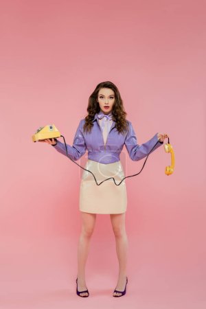 Puppen-Pose, junge Frau mit welligem Haar, Hörer und Retro-Telefon in der Hand, trendiges Outfit, brünettes Model in lila Jacke posiert und blickt in die Kamera auf rosa Hintergrund, Studioaufnahme 
