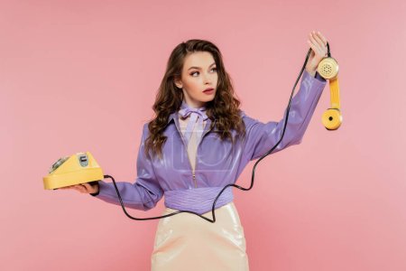 pose muñeca, hermosa mujer joven con el pelo ondulado mirando el teléfono mientras sostiene el teléfono retro amarillo, modelo morena en chaqueta púrpura posando sobre fondo rosa, tiro al estudio 