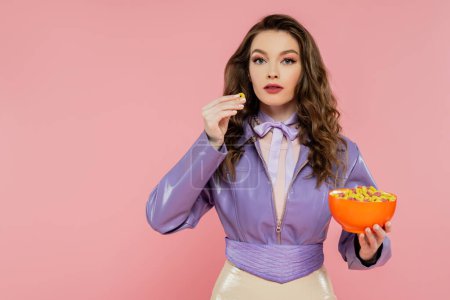 Konzeptfotografie, brünette Frau mit welligem Haar, die vorgibt, eine Puppe zu sein, Schüssel mit Cornflakes in der Hand, schnelles Frühstück, posiert auf rosa Hintergrund, stylische lila Jacke