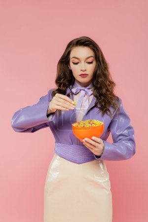 Konzeptfotografie, brünette Frau mit welligem Haar, die vorgibt, eine Puppe zu sein, Schale mit Cornflakes in der Hand, beim Frühstück, posiert auf rosa Hintergrund, trendige lila Jacke