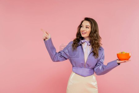 fotografía conceptual, mujer alegre con el pelo ondulado fingiendo ser una muñeca, apuntando hacia otro lado, sosteniendo tazón con hojuelas de maíz, comiendo sabroso desayuno, posando sobre fondo rosa, elegante chaqueta púrpura