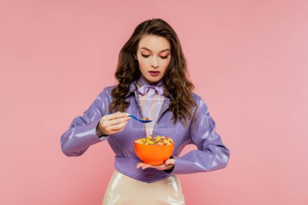 concept photography, brunette woman with wavy hair pretending to be a doll, holding bowl with corn flakes and spoon, eating tasty breakfast, posing on pink background, stylish purple jacket