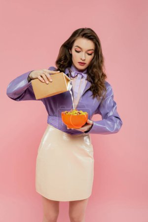 concepto de fotografía, muñeca como, mujer joven con el pelo ondulado celebración bowl con hojuelas de maíz, verter la leche de la caja de cartón, delicioso desayuno, posando sobre fondo rosa, elegante chaqueta púrpura