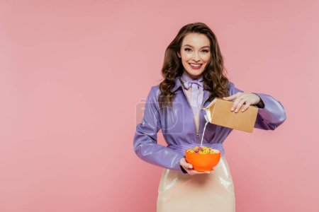 concepto de fotografía, muñeca como, mujer feliz con el pelo ondulado celebración bowl con hojuelas de maíz, verter la leche de la caja de cartón, sabroso desayuno, posando sobre fondo rosa, elegante chaqueta púrpura
