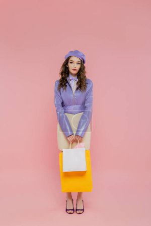 Foto de Consumismo, fotografía de moda, atractiva joven en boina sosteniendo bolsas de compras sobre fondo rosa, posando como una muñeca, de pie y mirando a la cámara, traje de moda, consumismo - Imagen libre de derechos