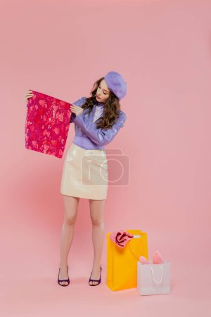 mode et consumérisme, attrayant jeune femme en béret tenant des vêtements de couleur vibrante près des sacs à provisions sur fond rose, consumérisme, debout dans une veste et une jupe en cuir à la mode, pleine longueur