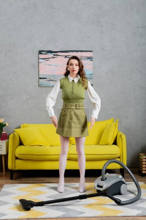 housekeeping concept, young woman with wavy hair standing on clean carpet near vacuum cleaner and yellow couch, gesturing and looking at camera, housewife in dress, domestic life, posing like a doll 
