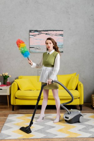 Photo for Housekeeping concept, young woman with wavy hair standing on carpet and holding dust brush, using vacuum cleaner, housewife in dress and white tights, domestic life, posing like a doll - Royalty Free Image