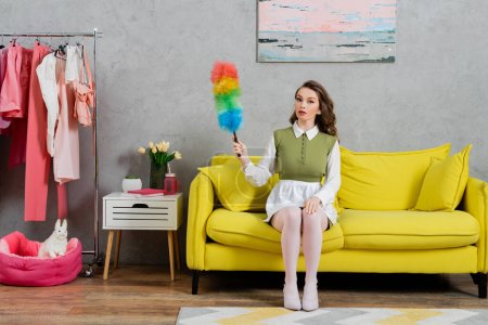Foto de Concepto de limpieza, mujer joven con el pelo ondulado sentado en el sofá y la celebración de cepillo de polvo, ama de casa en vestido y medias blancas mirando a la cámara, la vida doméstica, posando como una muñeca - Imagen libre de derechos