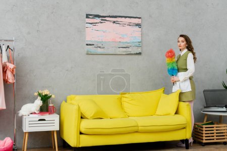 housekeeping concept, young woman with brunette wavy hair holding dust brush, housewife doing her daily duties, lifestyle, domestic chores, standing and looking at camera, painting on wall 