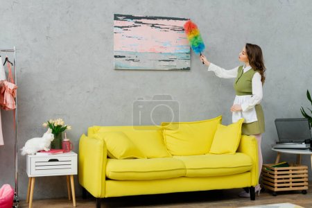 Photo for Housekeeping concept, young woman with brunette wavy hair cleaning dust off a painting, holding dust brush, housewife doing her daily duties, lifestyle, domestic chores - Royalty Free Image