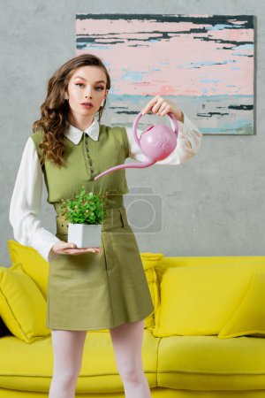 umweltfreundliches, hauswirtschaftliches Konzept, junge Frau mit welligem Haar, die grüne Pflanze wässert, Wachstum und Botanik, Hauswirtschaft, schöne Hausfrau, die in die Kamera schaut und im Wohnzimmer steht 