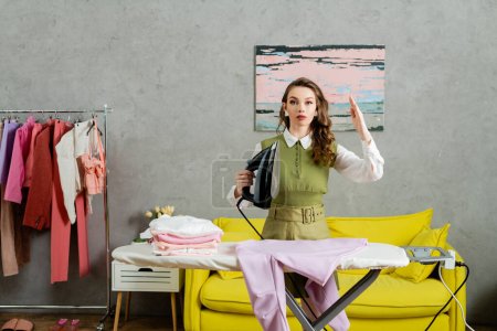 young woman with wavy hair acting like a doll, gesturing unnaturally, housewife holding iron near clean clothes and looking at camera, housekeeping concept, laundry day, daily routine 