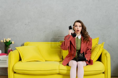 phone call, attractive woman with wavy hair sitting on yellow couch, housewife talking on retro telephone, acting like a doll, looking at camera with opened mouth, shock, grey wall 