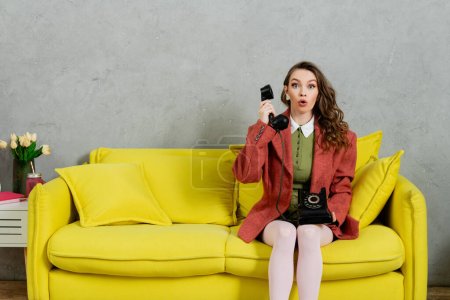 phone call, astonished woman with wavy hair sitting on yellow couch, housewife talking on retro telephone, acting like a doll, looking at camera with opened mouth, shock, grey wall 