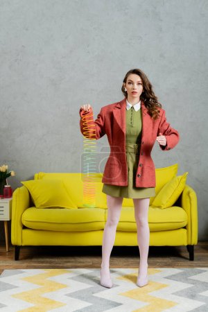 schöne Frau posiert wie eine Puppe und spielt mit Regenbogen slinky, Blick auf Kamera, modernes Wohnzimmer mit gelber Couch, kindisch, Vintage, Nostalgie, buntes Spielzeug, Freizeit und Spaß 