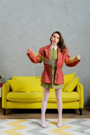 jeune femme brune posant comme une poupée et jouant avec arc-en-ciel slinky, regardant la caméra, salon moderne avec canapé jaune, enfantin, vintage, nostalgie, jouet coloré, loisirs et plaisir 