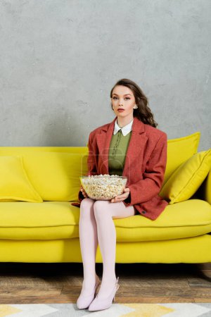 Konzeptfotografie, junge Frau mit brünetten welligen Haaren hält Schüssel mit Popcorn, salzigen Filmsnack, Home Entertainment, sitzt auf bequemem gelben Sofa und blickt in die Kamera
