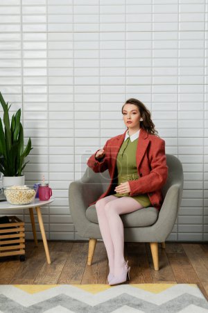 concept photographie, jeune femme aux cheveux bruns ondulés assis sur un fauteuil confortable à côté d'un bol avec du pop-corn, agissant comme une poupée et gesticulant anormalement, collation de film, regardant la caméra 
