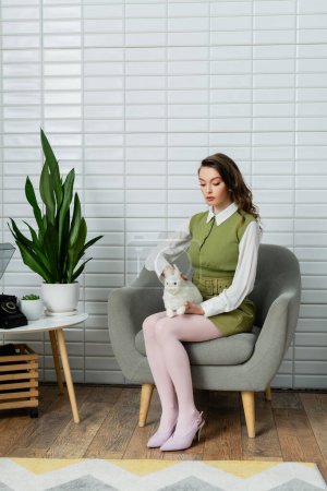 woman acting like a doll, beautiful woman sitting on comfortable grey armchair and holding toy rabbit, green plants and retro telephone on table, concept photography 