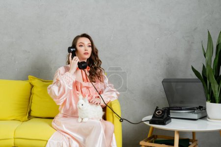 Vintage-Vibes, schöne Frau mit brünetten und welligen Haaren sitzt auf gelbem Sofa, Hausfrau spricht am Retro-Telefon, posiert wie eine Puppe, schaut weg, Plattenspieler 