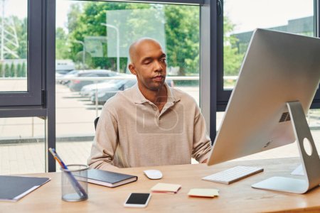 inclusion, bold african american man with myasthenia gravis, dark skinned office worker sitting at desk and using computer, looking at monitor, keyboard and mouse, smartphone with blank screen 