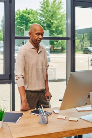syndrome de myasthénie grave, homme d'affaires afro-américain audacieux debout avec canne à pied et regardant l'écran d'ordinateur, employé de bureau à la peau foncée en tenue décontractée debout au bureau, inclusion 