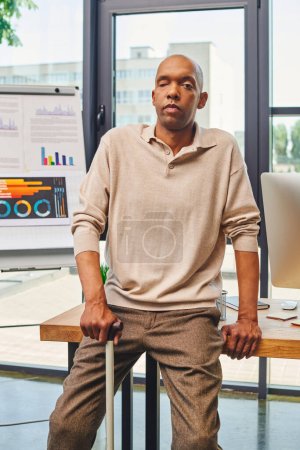 Inklusion, Myasthenia-Gravis-Syndrom, mutiger afrikanisch-amerikanischer Geschäftsmann, der mit Gehstock neben Computermonitor am Schreibtisch steht, dunkelhäutige Büroangestellte in Freizeitkleidung