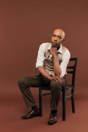 inclusion, bold african american man with myasthenia gravis syndrome, sitting on chair and looking at camera, dark skinned man with chronic disease on brown background