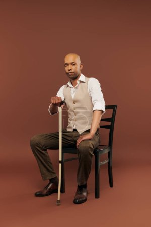 inclusion, bold african american man with myasthenia gravis syndrome, sitting on chair and leaning on walking stick, looking at camera, dark skinned man with chronic disease on brown background