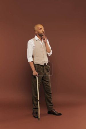 inclusion, thoughtful african american man with myasthenia gravis syndrome leaning on walking cane, looking away, bold dark skinned man with chronic disease standing on brown background 