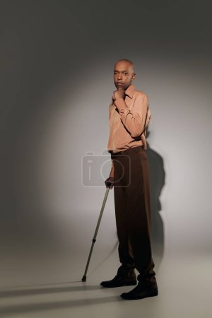 fetter afrikanisch-amerikanischer Mann mit Myasthenia-Gravis-Syndrom, stehend mit Gehstock auf grauem Hintergrund, dunkelhäutige Person im Hemd, Vielfalt und Inklusion, in die Kamera schauend, in voller Länge 
