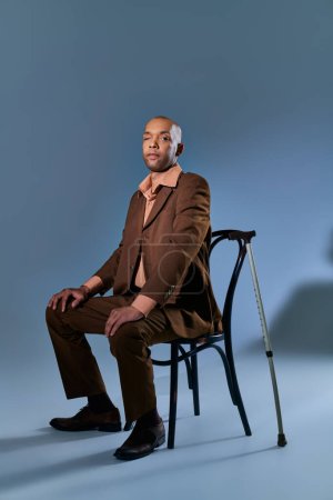 Vielfalt und Inklusion, afrikanisch-amerikanischer Mann mit Myasthenia-Gravis-Syndrom sitzt auf Stuhl und blickt in die Kamera auf blauem Hintergrund, Gehstock, Gehschwierigkeiten, dunkelhäutige Person im Anzug