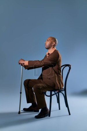 Vielfalt und Inklusion, körperliche Beeinträchtigung, afrikanisch-amerikanischer Mann mit Myasthenia gravis sitzt auf Stuhl und schaut vor blauem Hintergrund weg, lehnt sich an Gehstock, Schwierigkeiten beim Gehen